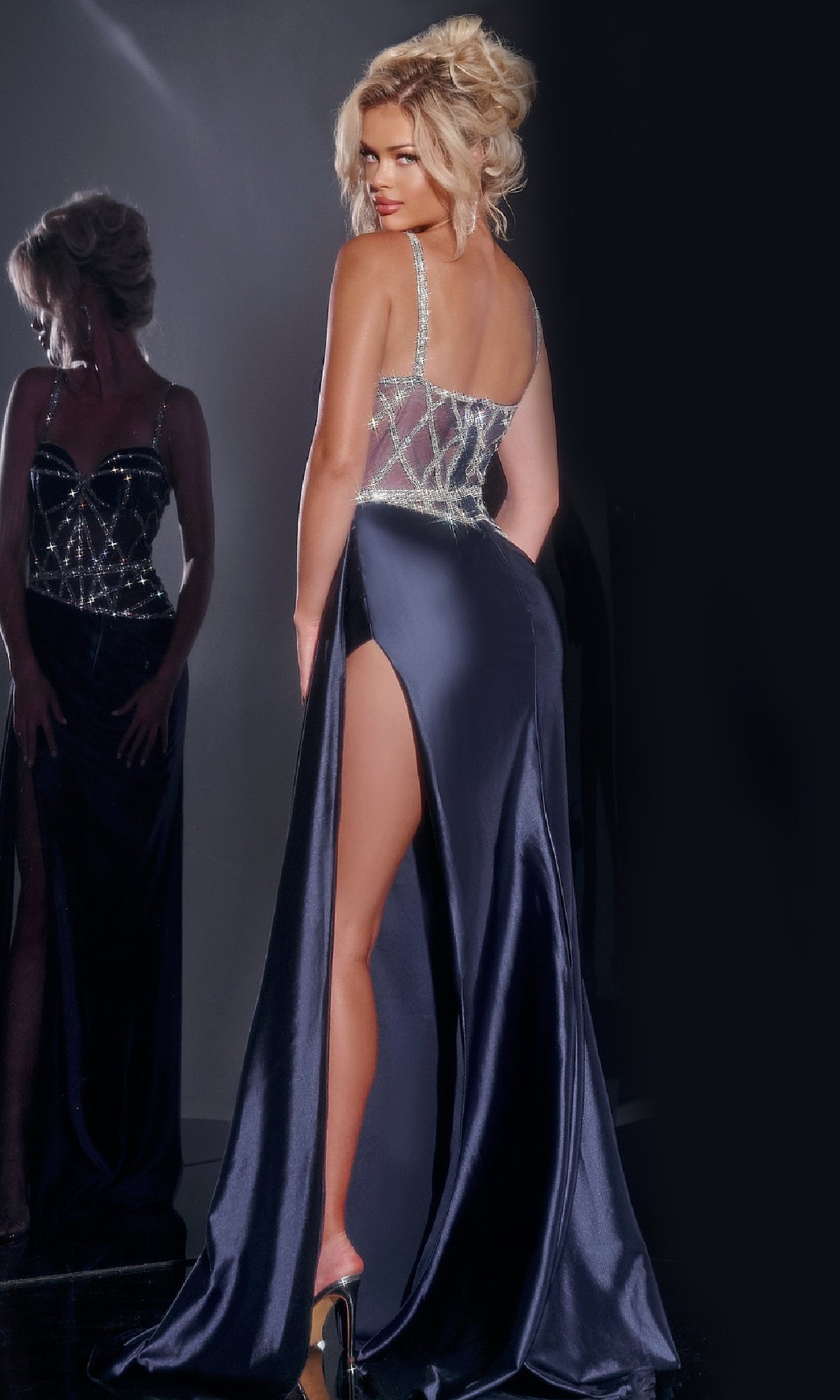 A woman poses with her back to the camera in front of a mirror, wearing the Jovani 40506 long prom dress—navy blue, form-fitting, with a high slit and sheer beaded bodice.